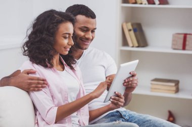 Tablet kullanma ve odada bir kanepede otururken gülümseyen genç güzel Afro-Amerikan Çift.