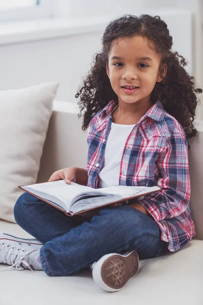 可爱的小黑人女孩 休闲服装 拿着一本书 坐在房间里的沙发上看着相机 — 图库照片