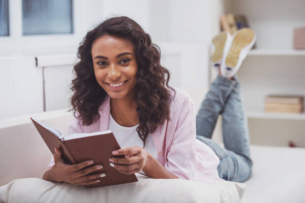 Young beautiful Afro-American woman reading a book, looking at camera and smiling while lying on a sofa in the room.