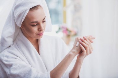 Ellerini tuvalette ayakta, krem uygulayarak başında bir havlu ile bornoz güzel genç kadın