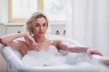 Köpük ile keyifli banyo keyfi, kameraya bakıyor ve gülümseyen güzel sarışın genç kadın