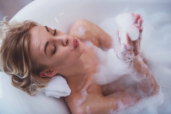 Top view of beautiful young blonde woman enjoying pleasant bath and blowing foam