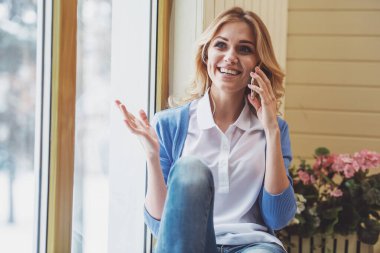 Mobil telefonda konuşurken, uzağa arıyorsunuz ve pencere pervazına üzerinde otururken gülümseyen güzel genç kadın