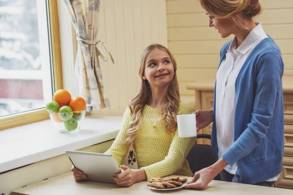 可爱的少女使用平板电脑 美丽的母亲抱着一个杯子和盘子里的饼干 看着对方和微笑 — 图库照片