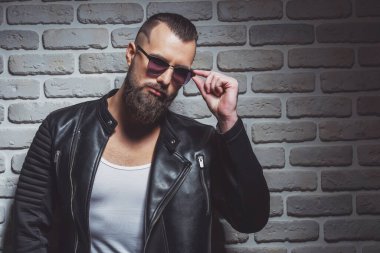 Çekici şık erkek deri ceket ve güneş gözlüğü kamera, duvara karşı ayakta bakarak sakallı