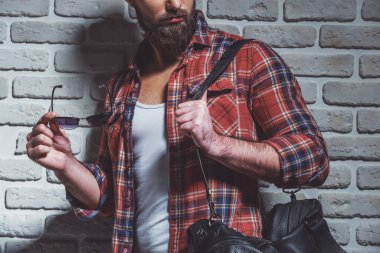 Bir siyah deri çanta ve güneş gözlüğü, kırpılmış tuğla duvara karşı ayakta tutan rahat gömlekli sakallı şık adam