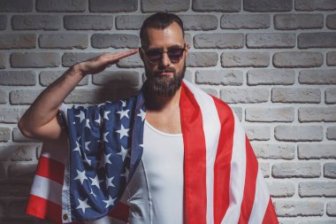 Kameraya bakarak ve selamlayan, Amerikan bayrağı duvara karşı duran sakallı şık adam kaplı
