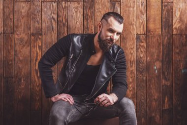 Güneş gözlüğü tutan ve kameraya bakan deri ceket sakallı şık adam, ahşap bir arka plan üzerinde oturan