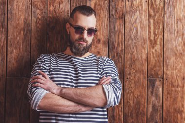 Çizgili yelek ve güneş gözlüğü sakallı şık adam, kameraya bakarak, ahşap bir arka plan üzerinde duran