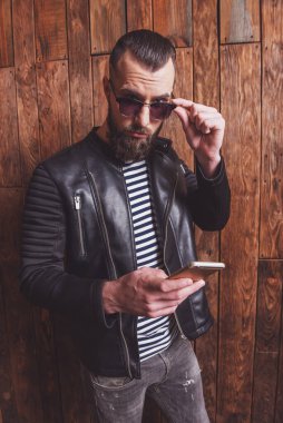 Bir cep telefonu kullanarak deri ceket ve güneş gözlüğü sakallı şık adam, ahşap bir arka plan üzerinde duran