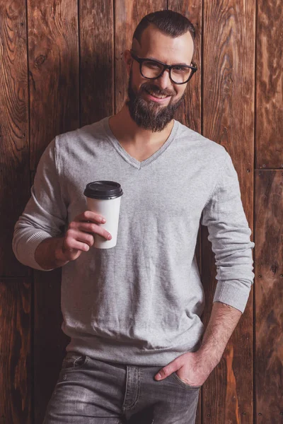 时髦的男子 留着灰色毛衣 戴着眼镜 拿着杯子 面带微笑 站在木制背景上 — 图库照片