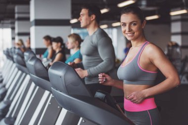 Spor salonunda bir koşu bandı üzerinde çalışan ve gülümseyerek kameraya bakarak çekici genç kadın