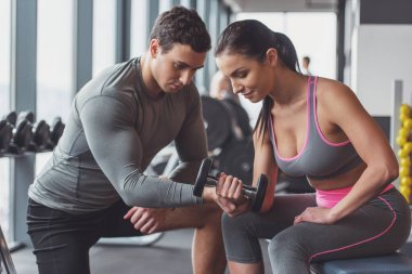 Spor salonunda, ona yardım yakışıklı kaslı eğitmen dumbbells ile çalışma dışarı çekici genç kadın