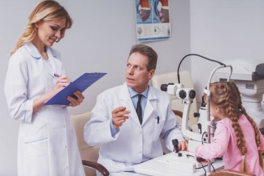 Küçük kızın gözleri modern ekipmanları ile inceleyerek yakışıklı göz doktoru. Güzel hemşire notlar