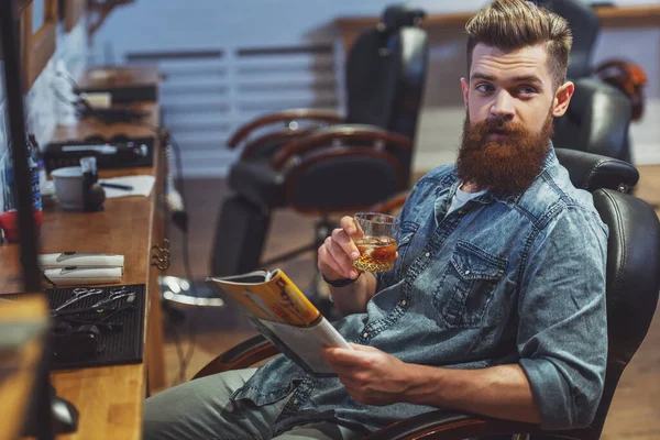 英俊的年轻人有胡子的人读一本杂志和坐在理发店的椅子上时 按住一杯饮料 — 图库照片