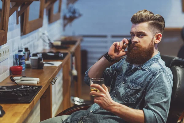 英俊的年轻人有胡子的人拿着一杯饮料 坐在理发店的椅子上同时在移动电话上交谈 — 图库照片