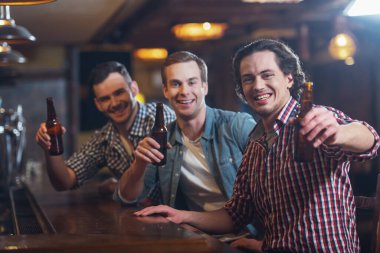Üç genç adam rahat kıyafetler içinde gülümseyen, kameraya bakıyor ve sayaç pub bar, otururken bira şişesi holding