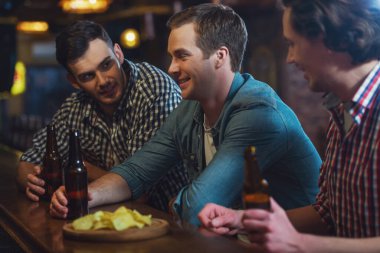 Üç genç adam rahat kıyafetler içinde konuşurken, cipsi yemek ve sayaç pub bar, otururken bira içmek