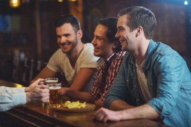 Üç genç adam rahat kıyafetler içinde bir barmenle konuşuyor, cipsi yemek ve sayaç pub bar, otururken bira içmek