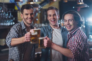 Three young men in casual clothes are smiling, looking at camera and clanging glasses of beer together while standing near bar counter in pub clipart