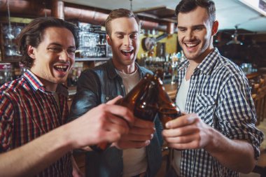 Üç genç adam rahat kıyafetler içinde gülümseyen ve bira şişesi birlikte sayaç pub bar yakınındaki dururken kımıldıyor.