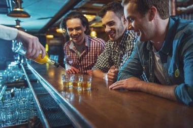 Üç genç adam rahat kıyafetler içinde sayaç barda, bir barmen dökme alkollü içecek gözlük içine otururken gülüyorsun