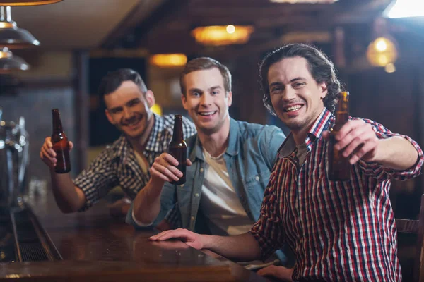 三名年轻男子穿着休闲服微笑着看着相机 坐在酒吧柜台在酒吧里拿瓶啤酒 — 图库照片