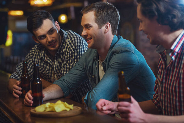 Three young men in casual clothes are talking, eating chips and drinking beer while sitting at bar counter in pub