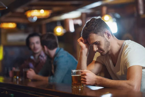 シャツで悲しそうな若い人は 人の男性バーのバック グラウンドのパブでカウンターに座ってビールのグラスを見てください — ストック写真