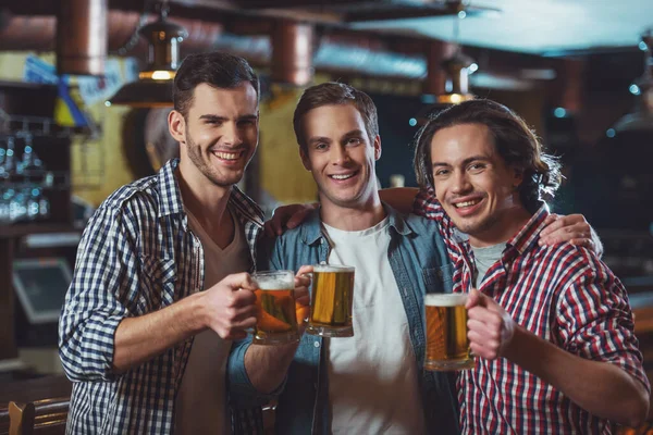 Trzech Młodych Mężczyzn Ubranie Uśmiechnięte Patrząc Kamery Trzymając Szklanki Piwa — Zdjęcie stockowe