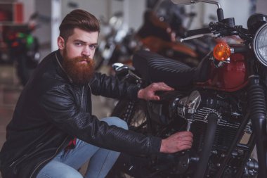Siyah deri ceketli yakışıklı genç sakallı adamı onun motosiklet incelenirken kameraya bakıyor