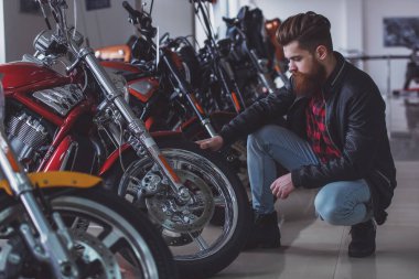 Sakallı yakışıklı genç siyah deri ceketli bir motosiklet salonda motosiklet inceleyerek