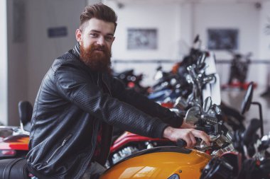 Yakışıklı genç sakallı adam kameraya bakarak ve bir motosiklet üzerinde otururken gülümseyen siyah deri ceketli yan görünüm