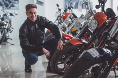 Siyah deri ceketli çekici genç sarışın adam kameraya bakıyor ve motosiklet incelenirken gülümseyen