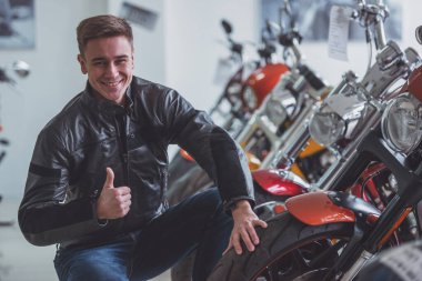 Siyah deri ceketli çekici genç sarışın adam kameraya bakarak, Ok işaret gösteren ve motosiklet incelenirken gülümseyen