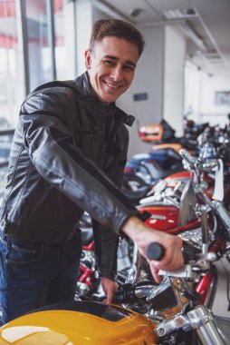 Siyah deri ceketli çekici genç sarışın adam kameraya bakıyor ve motosiklet bir salonda incelenirken gülümseyen