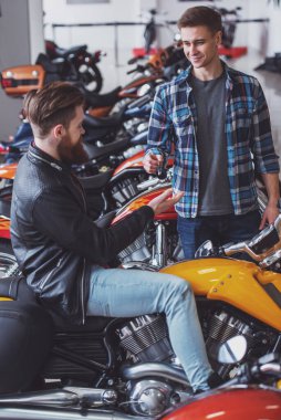 Sakallı yakışıklı genç siyah deri ceketli bir motosiklet satın alıyor, çekici sarışın adam anahtarlarını veriyor