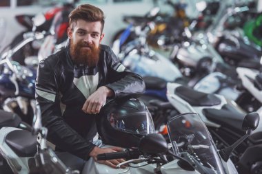 Sakallı yakışıklı genç siyah deri ceketli bir kask holding, kameraya bakıyor ve bir motosiklet üzerinde otururken gülümseyen