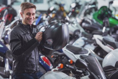 Çekici genç sarışın adam siyah deri ceketli bir kask holding, kameraya bakıyor ve bir motosiklet üzerinde otururken gülümseyen