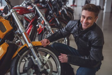 Siyah deri ceketli çekici genç sarışın adam kameraya bakıyor ve bir motosiklet incelenirken gülümseyen