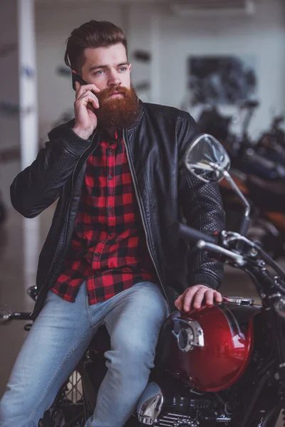 留胡子的英俊年轻人在黑色皮夹克是谈移动电话 坐在摩托车上视线 — 图库照片