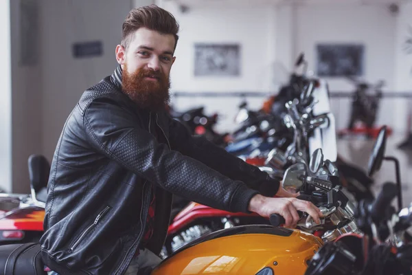 留胡子的英俊年轻人在看着相机 微笑着坐在一辆摩托车上的黑色皮夹克的侧视图 — 图库照片