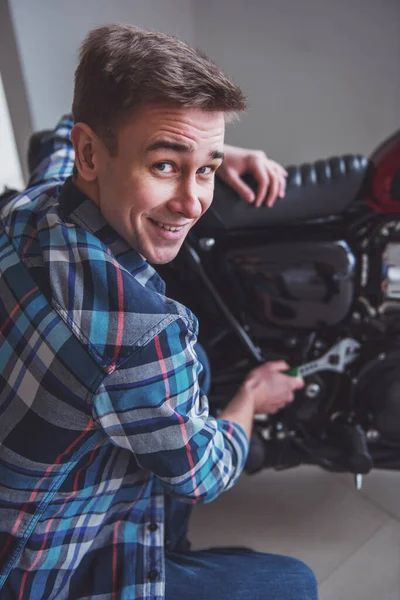 カジュアルな服で魅力的な金髪の男はカメラを見て バイクを修理しながら笑みを浮かべて — ストック写真