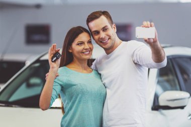 Güzel genç çift gülümseyen ve Otomobil Fuarı bir araba satın alma sırasında bir akıllı telefon kullanarak bir selfie yapma. Kadın anahtarları tutuyor