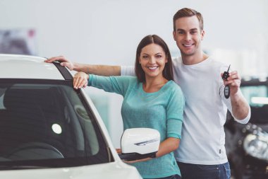 Güzel genç çift gülümseyen ve yeni arabalarında bir Otomobil Fuarı olarak yaslanmış iken kameraya bakıyor. Adam arabanın anahtarlarını tutuyor