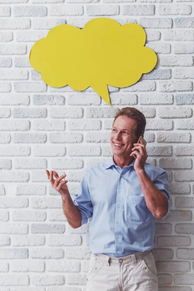 Handsome middle aged man is talking on the mobile phone and smiling while standing against white brick wall, yellow speech bubble above