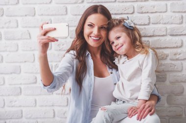 Güzel genç anne bir akıllı telefon kullanarak ve, beyaz tuğla duvara gülümseyen sevimli küçük kızıyla birlikte bir selfie yapıyor