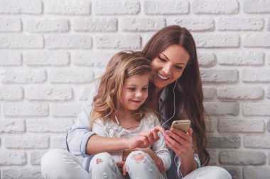 Güzel genç anne ve onun sevimli küçük kızı beyaz tuğla duvara karşı, akıllı telefon kullanarak müzik dinliyor ve gülümseyerek