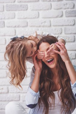 Güzel genç anne ve küçük kızı beyaz tuğla duvara karşı oturan, gülümsüyor. Kız annesinin gözlerini kapatıyor.