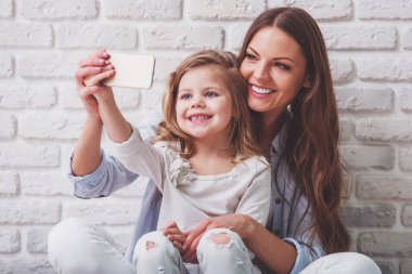 Güzel genç anne bir akıllı telefon kullanarak ve, beyaz tuğla duvara gülümseyen sevimli küçük kızıyla birlikte bir selfie yapıyor